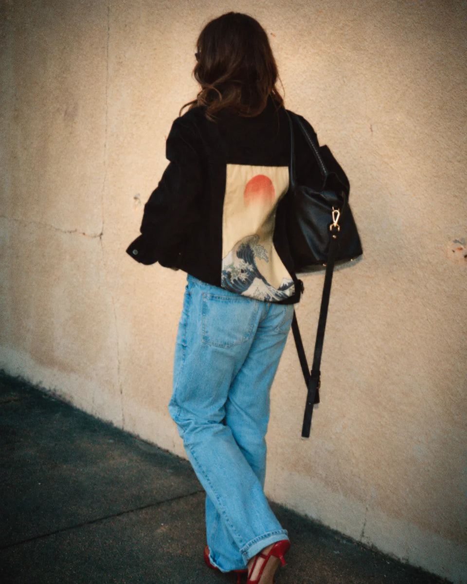 Person in a black jacket with a wave and red sun art panel, paired with blue jeans.
