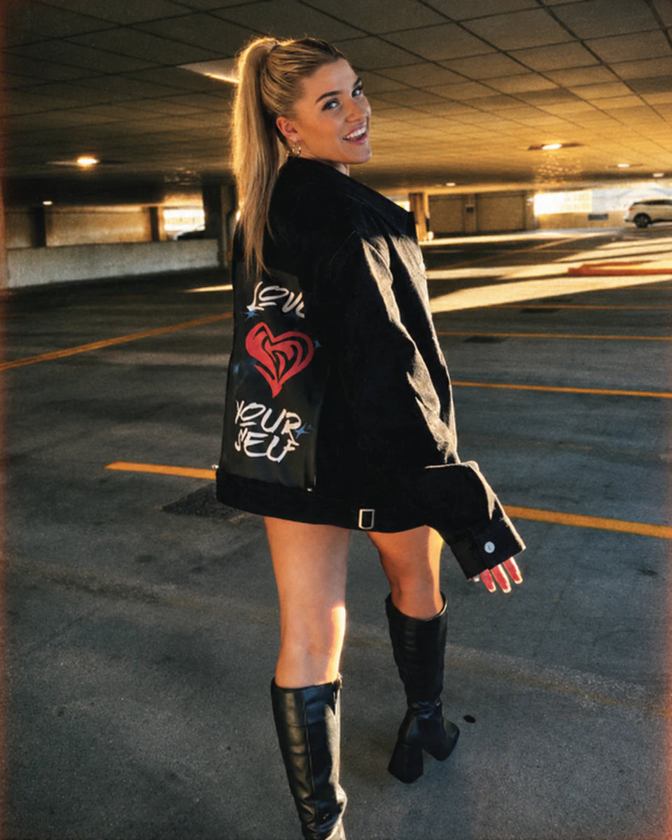 Person in a black jacket with a red heart art panel and tall black boots.