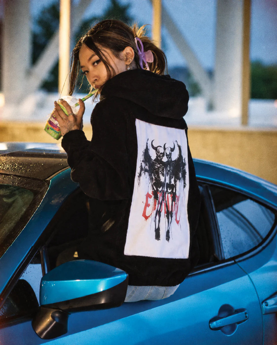 Person in a black hoodie with a skeletal figure art panel, leaning out of a blue car.
