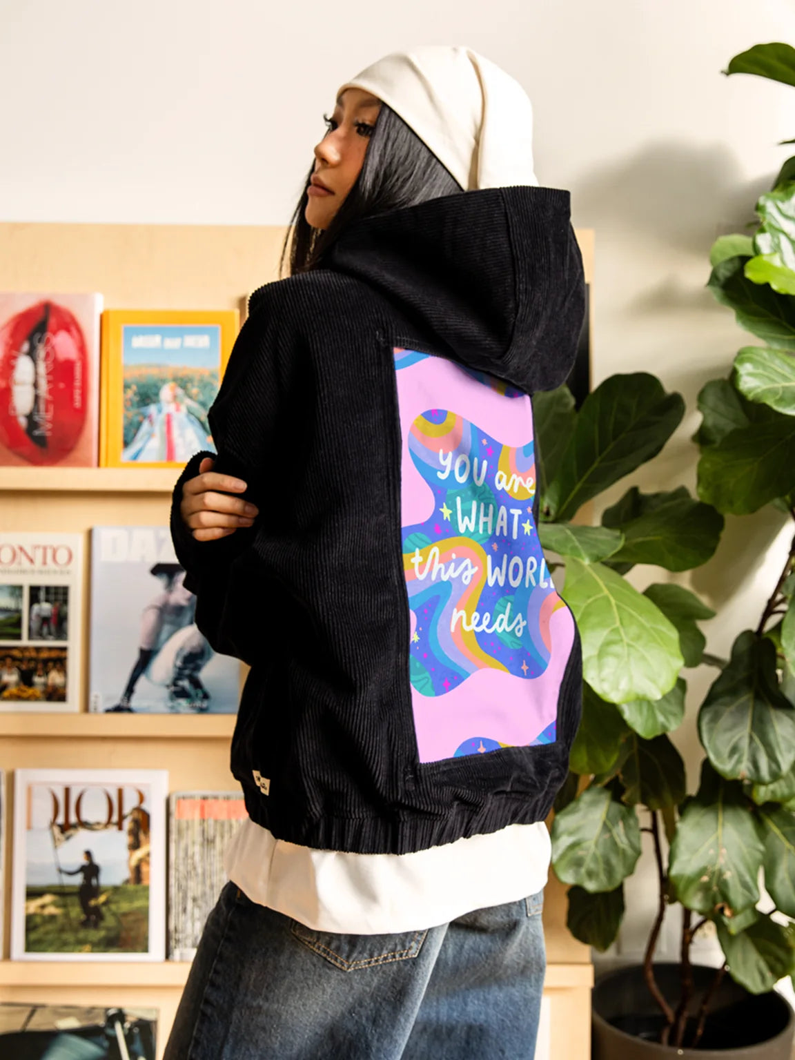 Woman wearing an Onyx corduroy hoodie with pink and blue inspirational back art in a book-filled studio.