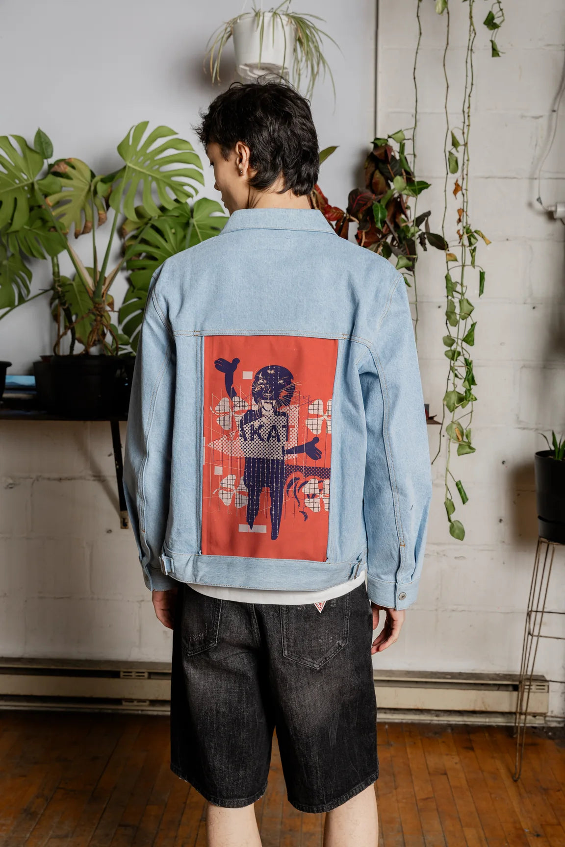 Man wearing an Oceanside denim jacket with red illustrated back art in a plant-filled studio.