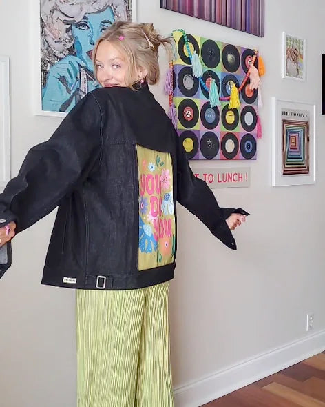Creator showing colorful sunshine back art on a Raven denim jacket with green trousers in a music-filled room.