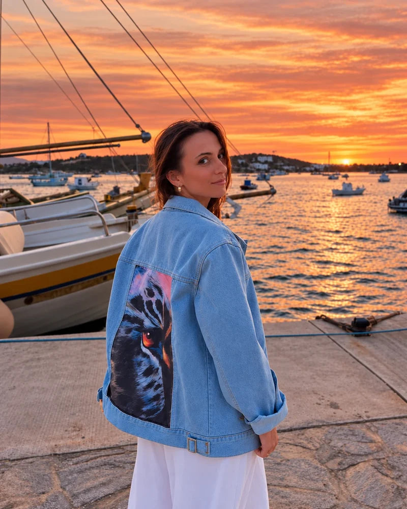 Customer wearing an Oceanside denim jacket with feather artwork beside a harbor at sunset.